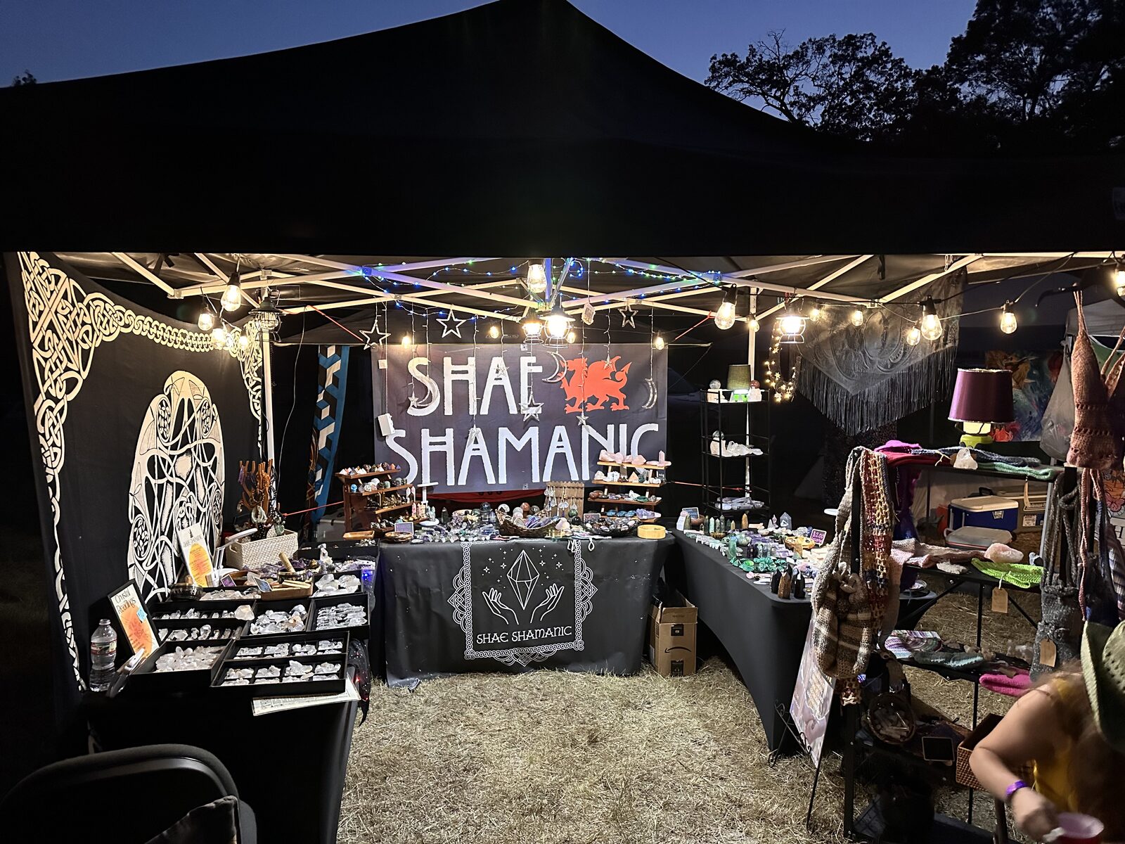 Shae Shamanics market booth at dusk, lit by string lights under a black canopy tent