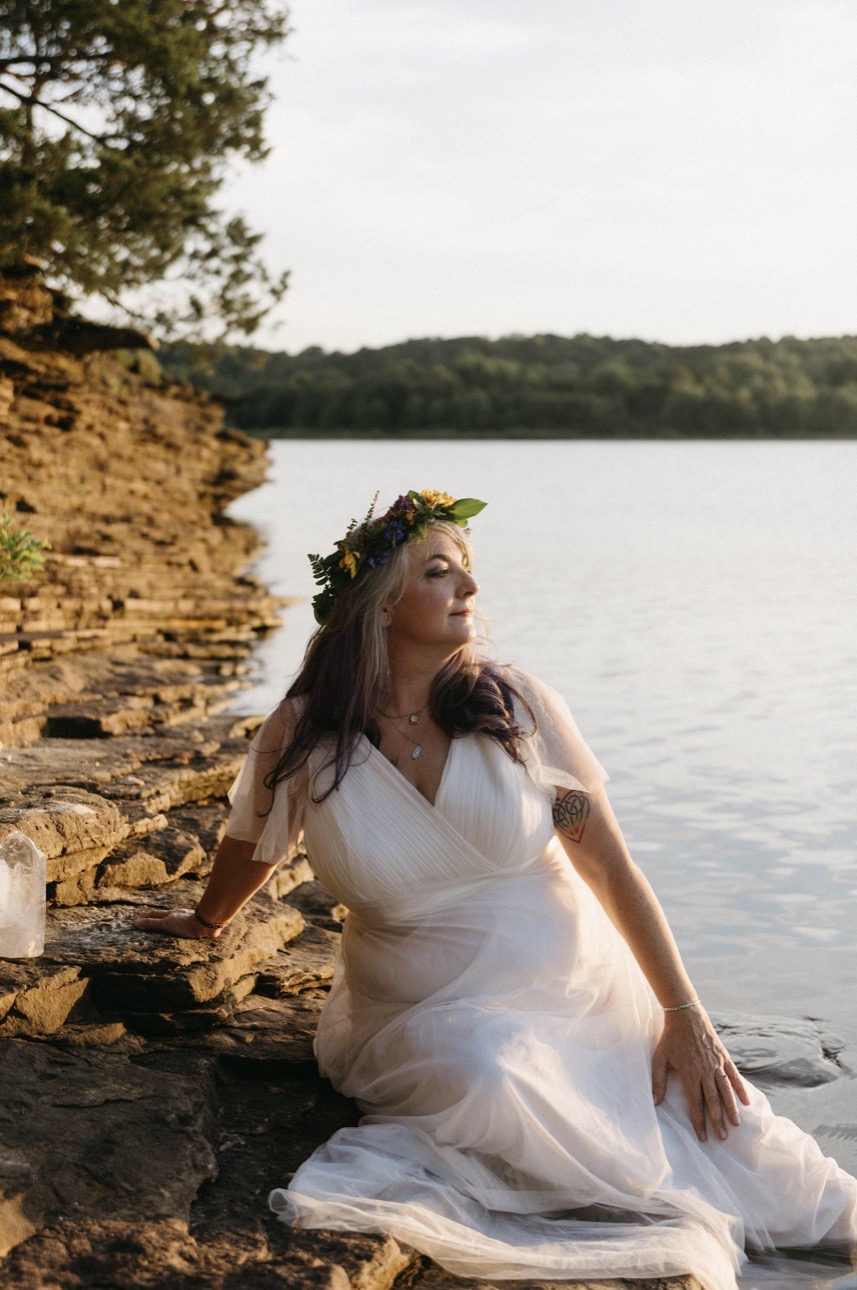 Shae seated on the rocks at the water's edge in a white dress and flower crown, a crystal beside her in the golden light